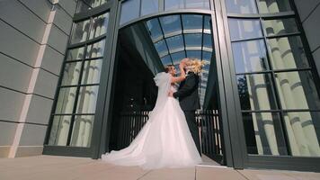 Joyful Bride and Groom Embracing Outside Modern Architecture, Bride in a flowing gown and groom in a suit embrace joyfully outside a contemporary building with large windows. video