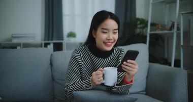 retrato de contento joven asiático mujer sentar en sofá disfrutar chateando en móvil teléfono mientras Bebiendo café en Mañana en vivo habitación, gratis tiempo, toma descanso, sonriendo foto