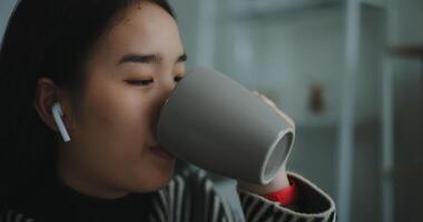 retrato de contento joven asiático mujer disfrutar escuchando a el música con inalámbrico auriculares mientras sentado bebida café en sofá en vivo habitación a hogar, ocio y estilo de vida, gratis hora foto