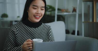 retrato de contento joven asiático mujer sentar en sofá Bebiendo café o té mientras trabajando creativo trabajo desde hogar en ordenador portátil con en línea red, tomar descanso, sonriendo foto