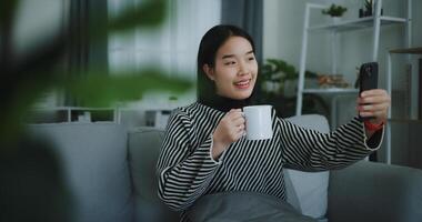 Portrait of Happy young asian woman sit on sofa holding coffee cup and enjoy taking selfies on mobile phone at home,Free time,take break,smiling photo