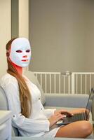 A beautiful girl with an LED mask on her head works at home on a laptop. Home skin care concept. photo