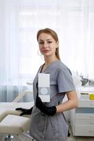 Happy female cosmetologist holding box with hands. Portrait pretty woman wearing black gloves and uniform, showing small box. Beauty, cosmetology concept. photo