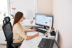 Portrait of a remote worker. A beautiful woman sits at a table in front of a computer and designs a building. Confident young lady freelancer working from home. photo