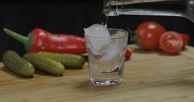 Pour vodka from a bottle into shot glass with ice cubes on the table photo