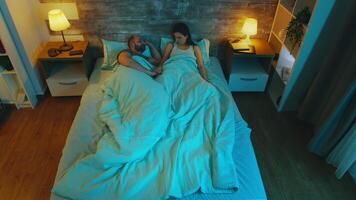 Top view of young couple having a conversation late at night in bed under the sheet with blue moon light in the room. video