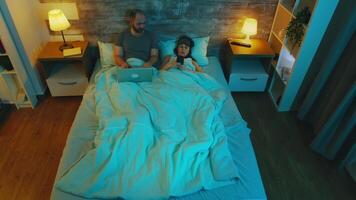 Top view of young couple under the sheets planning their vacation on laptop and smartphone. video