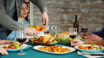 Zoom in shot of young man using sharp knife to slice roasted chicken at family dinner. video