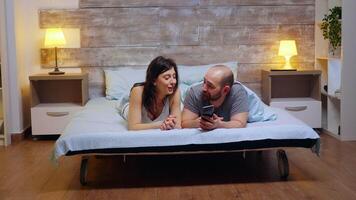 Ingrandisci nel tiro di di coppia nel pigiama sorridente mentre utilizzando smartphone nel letto. video