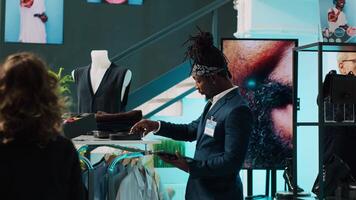 African american worker managing clothes stock on hangers, working on inventory using tablet in shopping center. Male store assistant in a suit preparing fashion collection on display. Camera A. video