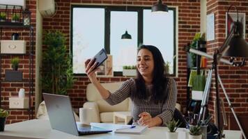 lachend gelukkig Indisch vrouw nemen selfie met mobiel telefoon terwijl glimlachen Bij huis kantoor bureau. persoon in mooi hoor elegant modern huis vastleggen foto gebruik makend van smartphone camera, camera een video