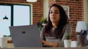 Porträt von heiter Freiberufler Überprüfung ihr E-Mails auf Notizbuch Gerät während beim Computer Schreibtisch. selbst beschäftigt Frau Eingabe Daten auf Laptop im hell gemütlich Wohnung Büro, lösen Aufgaben video