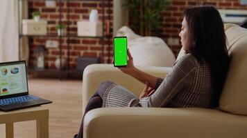 Remote worker looking at isolated screen cellphone while optimizing market performance charts. Indian woman checking online business analytics statistical data metrics on chroma key phone and laptop video