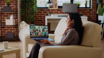 Teleworking team leader sitting on couch in cozy apartment, chatting with coworkers during teleconference meeting. Indian business manager at home holding online videocall with workers, camera B video