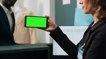 Store worker using smartphone with greenscreen template at cash register, working on commercial activity with male customer. Female employee looks at chromakey layout and copyspace. Camera A. video