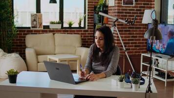 avlägsen anställd på skrivbord håller på med göra flera saker samtidigt, korskontroll data mellan bärbar dator och anteckningsblock. indisk kvinna i lägenhet påfrestande till komplett jobb uppgifter innan deadline, analyserar info, kamera en video