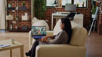 Teleworking team leader sitting on couch in cozy apartment, chatting with coworkers during teleconference meeting. Indian business manager at home holding online videocall with workers, camera A video