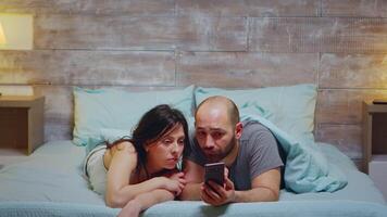 Husband and wife using phone while laying in bed wearing pajamas. video