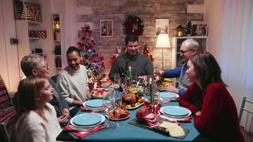 joven hombre tintinamente un vaso de champán con su padre celebrando Navidad. tradicional festivo Navidad cena en multigeneracional familia. disfrutando Navidad comida banquete en decorado habitación. grande familia reunión video