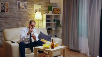 Businessman sitting on couch eating takeaway noodles from box after work in his apartment watching tv. video