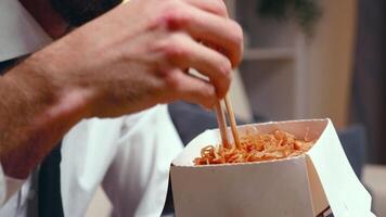 vicino su di stanco imprenditore mangiare tagliatelle con bastoncini nel vivente camera mentre Guardando tv. video