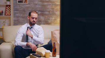 caucasian affärsman lossa hans slips Sammanträde på soffa efter en lång dag på arbete. äta hamburgare. video