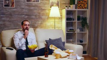 Businessman sitting on couch eating chips eating television. Zoom in shot. video