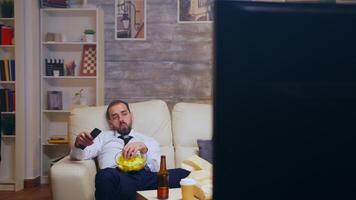 Tired and bored businessman with tie relaxing watching tv eating chips using remote control. video