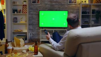 Back view of young man sitting on couch in front of tv with green screen using remote control. video