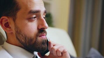 Close up of young businessman having a conversation on mobile phone sitting on couch in his apartment while watching tv. video