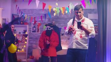 gespenstisch Zombie mit ein Axt mit seine Telefon beim Halloween Party mit andere Menschen verkleidet wie Monster. video