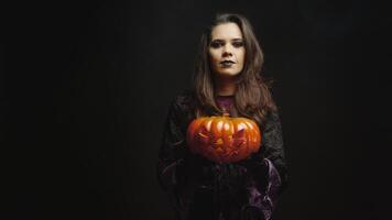 Young woman dressed up like a witch holding a pumpkin for halloween looking at the camera over a black background. video