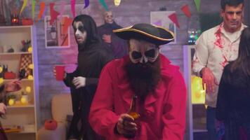 Handsome pirate with an axe drinking beer celebrating halloween with his scary friends dancing in decorated room. video
