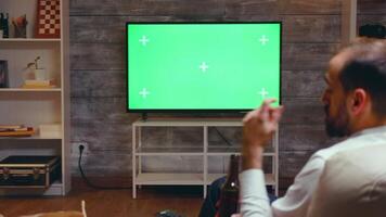 Back view of businessman sitting on couch eating chips drinking beer and looking at tv with green screen. video