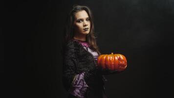 Beautiful young woman with evil face dressed up like a witch holding a pumpkin for halloween over a black background video