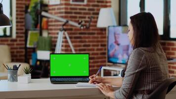 Teleworker staying at home using green screen laptop to do various job tasks at wooden desk. Remote working employee typing data on chroma key notebook keyboard and writing on notepad video