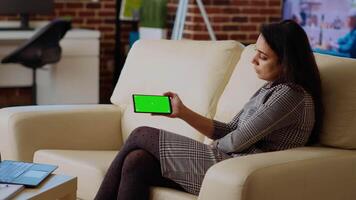 Relaxed stay at home worker enjoying herself, watching videos on chroma key smartphone. Chilled out woman sitting on couch looking at internet clips on isolated screen phone