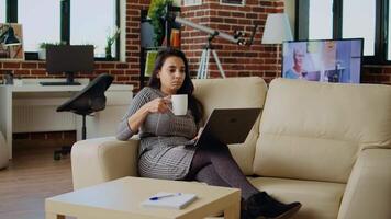 teletrabalhador deitado em sofá, bebendo café enquanto remotamente realizando tarefas dentro pessoal escritório. trabalhador autonomo sentindo-me estressado, desfrutando quente bebida para relaxar enquanto introduzindo dados em computador portátil, Câmera uma video