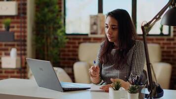 avlägsen anställd på skrivbord håller på med göra flera saker samtidigt, korskontroll data mellan bärbar dator och anteckningsblock. indisk kvinna i lägenhet påfrestande till komplett jobb uppgifter innan deadline, analyserar info, kamera b video