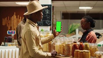 africain américain vendeur en portant téléphone intelligent app avec écran vert, en présentant isolé maquette modèle dans local supermarché. végétalien marchande les usages chromakey disposition à biologique épicerie magasin. caméra un. video