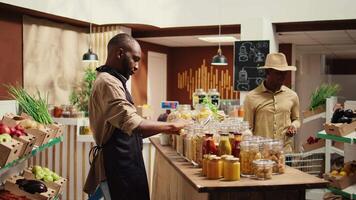 marchande avec tablier placement masse articles sur étagères à éco magasin, justificatif zéro déchets concept et durable mode de vie. africain américain vendeur La publicité fait maison garde-manger fournitures. caméra b. video