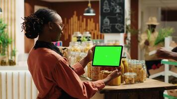 afrikansk amerikan kvinna användningar grönskärm på läsplatta i mataffär, stående Nästa till naturlig organisk Produkter. kvinna Säljare innehav grej som visar isolerat copy attrapp layout. kamera a. video