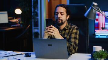 Man relaxing at home sitting at office desk, texting friends on smartphone in front of laptop. Cheerful freelancer taking break to chat with mates online using mobile phone in living room, camera A video