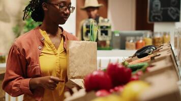 regular cliente colocando bio caseiro legumes dentro uma papel bolsa, indo mercearia compras às zero desperdício eco loja. mulher apoiando pequeno o negócio e sustentável estilo de vida. Câmera uma. portátil tomada. video