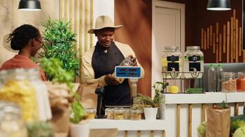 Merchant greeting woman at zero waste eco store checkout, weighting organic produce and bulk items in jars at cash register. African american client buying locally grown natural food. Camera A. video