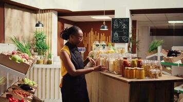 African american vendor arranging bulk products at local store, promoting zero waste concept for sustainable lifestyle. Merchant smiling and preparing to welcome clients at bio shop. Camera B. video