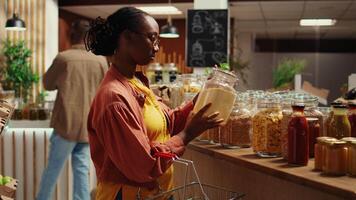 Female client checking variety of grains or pasta in reusable jars, looking to buy chemicals free pantry supplies from local farmers market. African american buyer shopping for bio items. Camera B. video