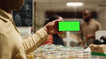 afrikanisch amerikanisch Verkäufer zeigt an grüner Bildschirm Anzeige auf Handy, Mobiltelefon Telefon im lokal Gegend Markt, Verkauf natürlich Bulk Produkte. Händler hält Gerät mit leer Copyspace Attrappe, Lehrmodell, Simulation Bildschirm. Kamera a. video