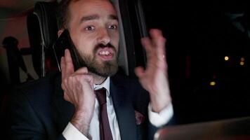 Bearded businessman in business suit having a phone call while working on laptop from the back seat of his limousine with personal driver. video