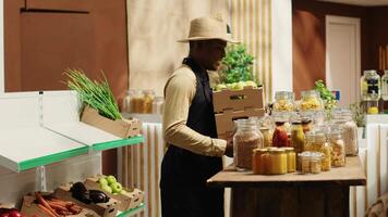 Local vendor placing crates with freshly harvested fruits on grocery store display, recommending organic produce to regular customer. African american farmer restocking all merchandise. Camera A. video